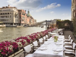 Un'area pranzo in riva al fiume con tavoli apparecchiati per un pasto, affacciata su un canale con edifici storici e un ponte.
