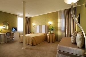 Una camera d'albergo con un letto, una poltrona letto, una scrivania con televisione e una pianta in vaso.