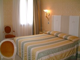 Una camera d'albergo con due letti, una sedia, un tavolo e una lampada a parete.