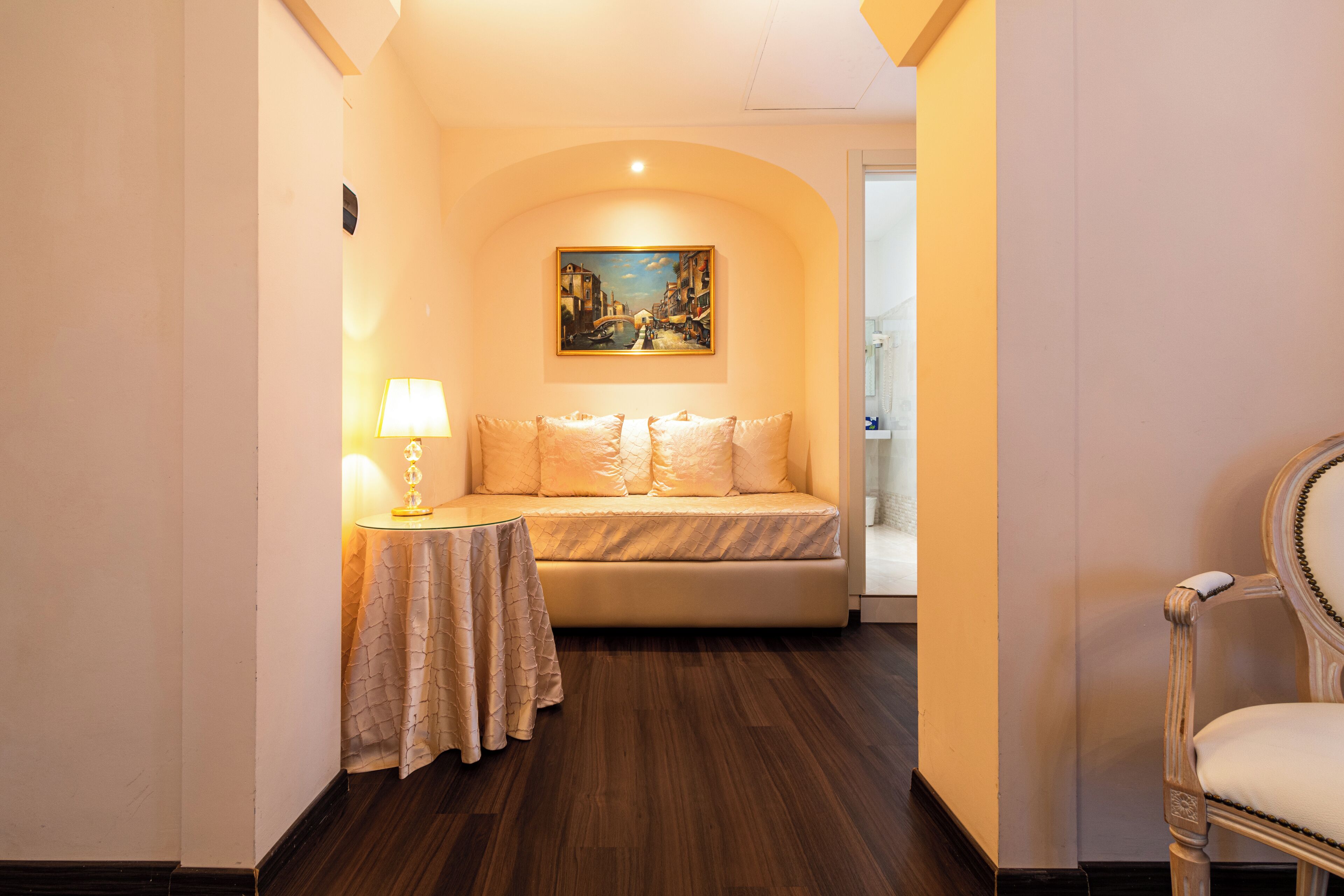 A hotel room with a sofa, a table with a lamp, a painting on the wall, and a chair.