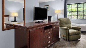 A hotel room with a wooden cabinet, a television, a green armchair, and a window with a view of greenery. - Days Inn by Wyndham Iron Mountain (Iron Mountain)