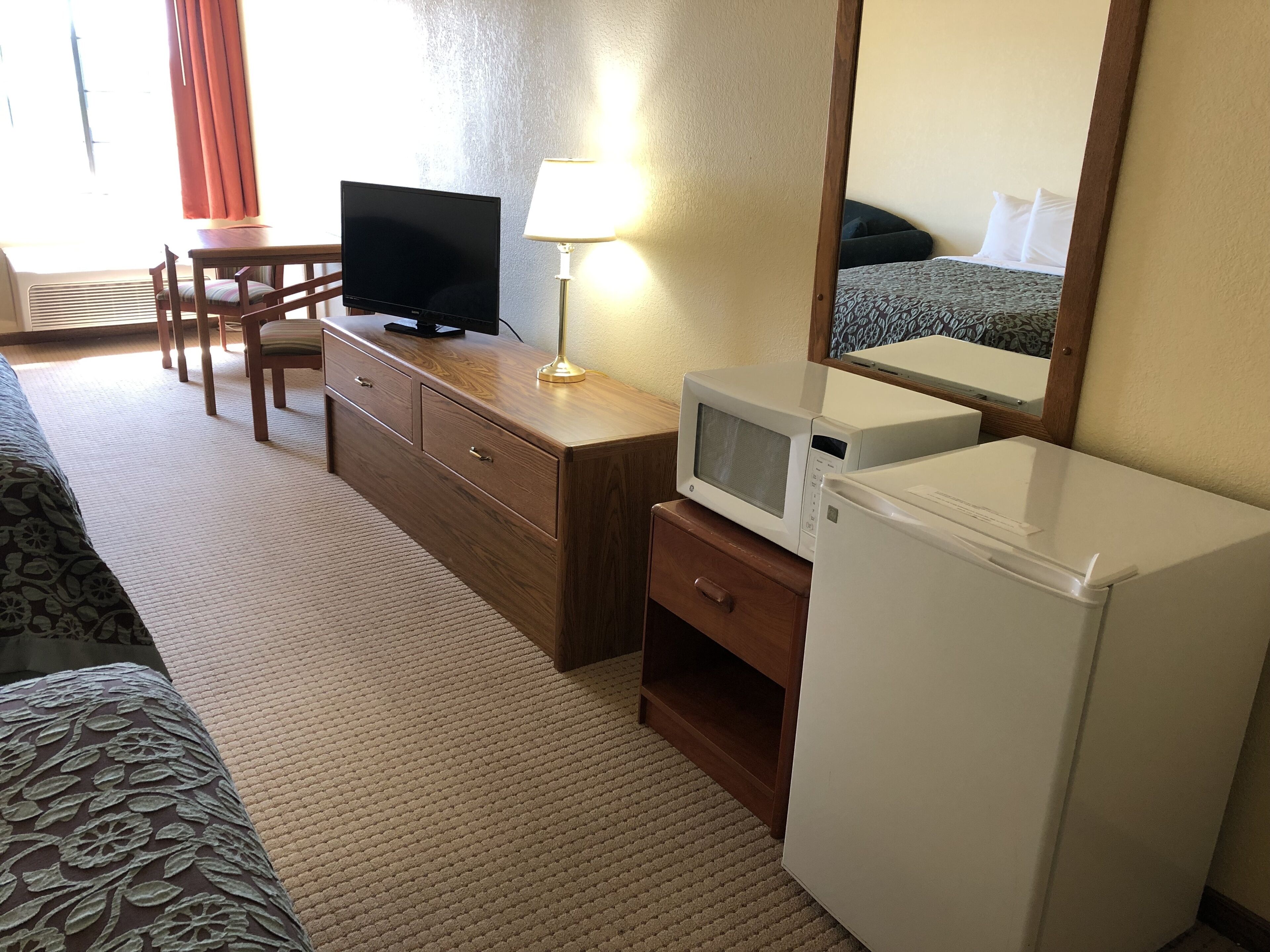 A hotel room with a wooden desk, a television, a microwave, a refrigerator, a bed, and a mirror.