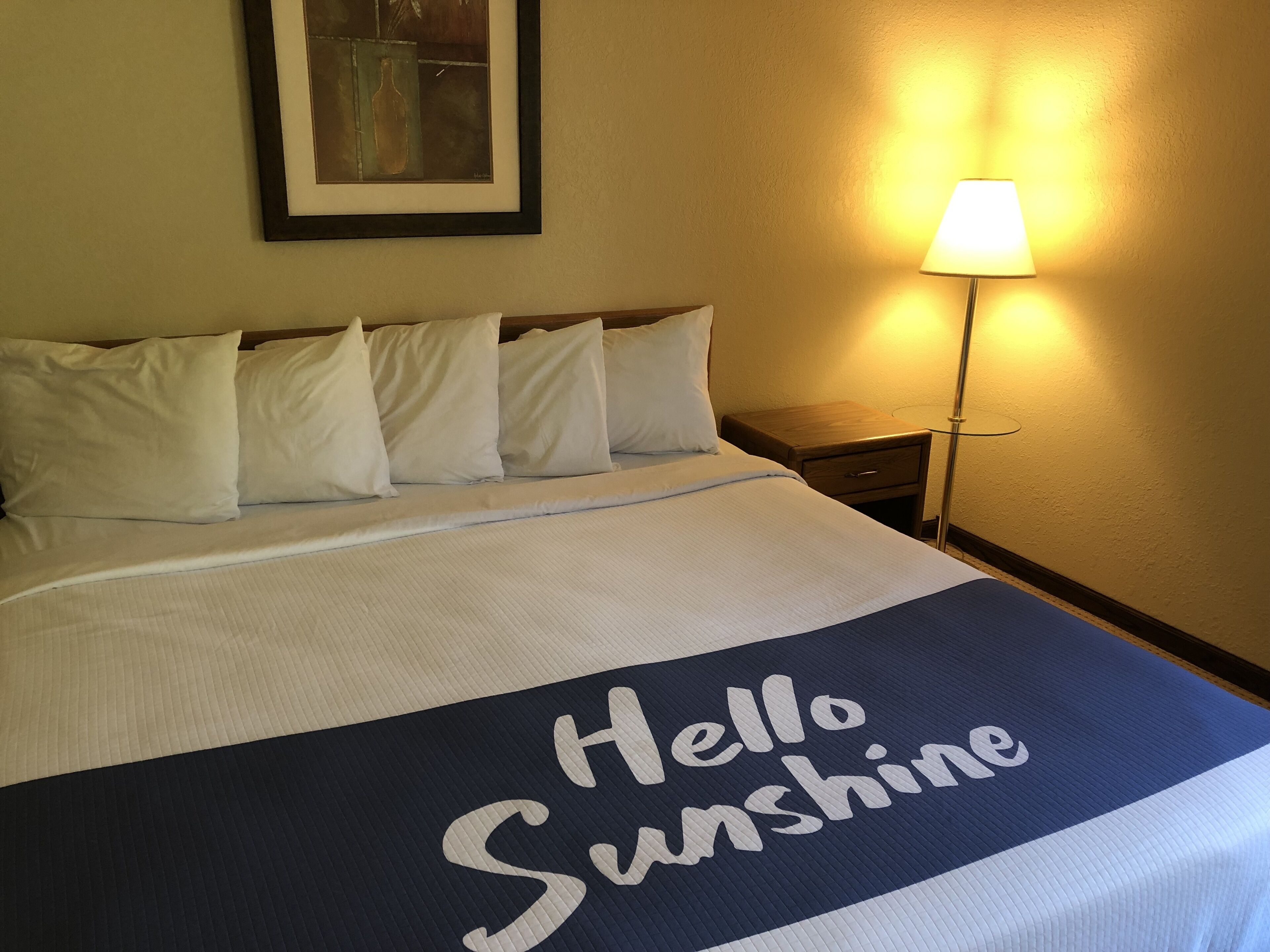 A neatly made bed with a "Hello Sunshine" sign, a framed artwork on the wall, and a bedside lamp.