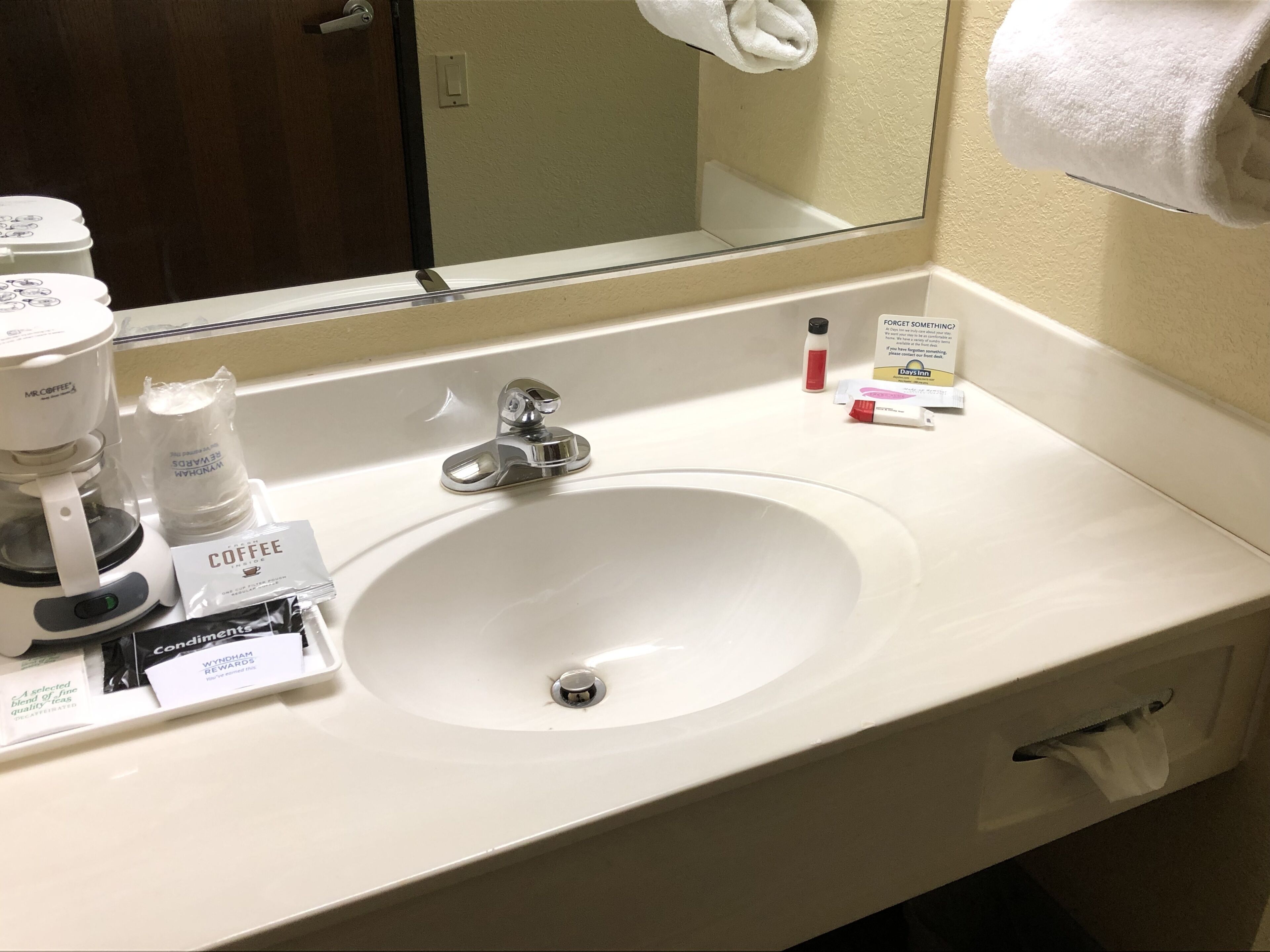 Un baño de hotel con un lavamanos blanco, un espejo, una toalla colgada en la pared y una cafetera.