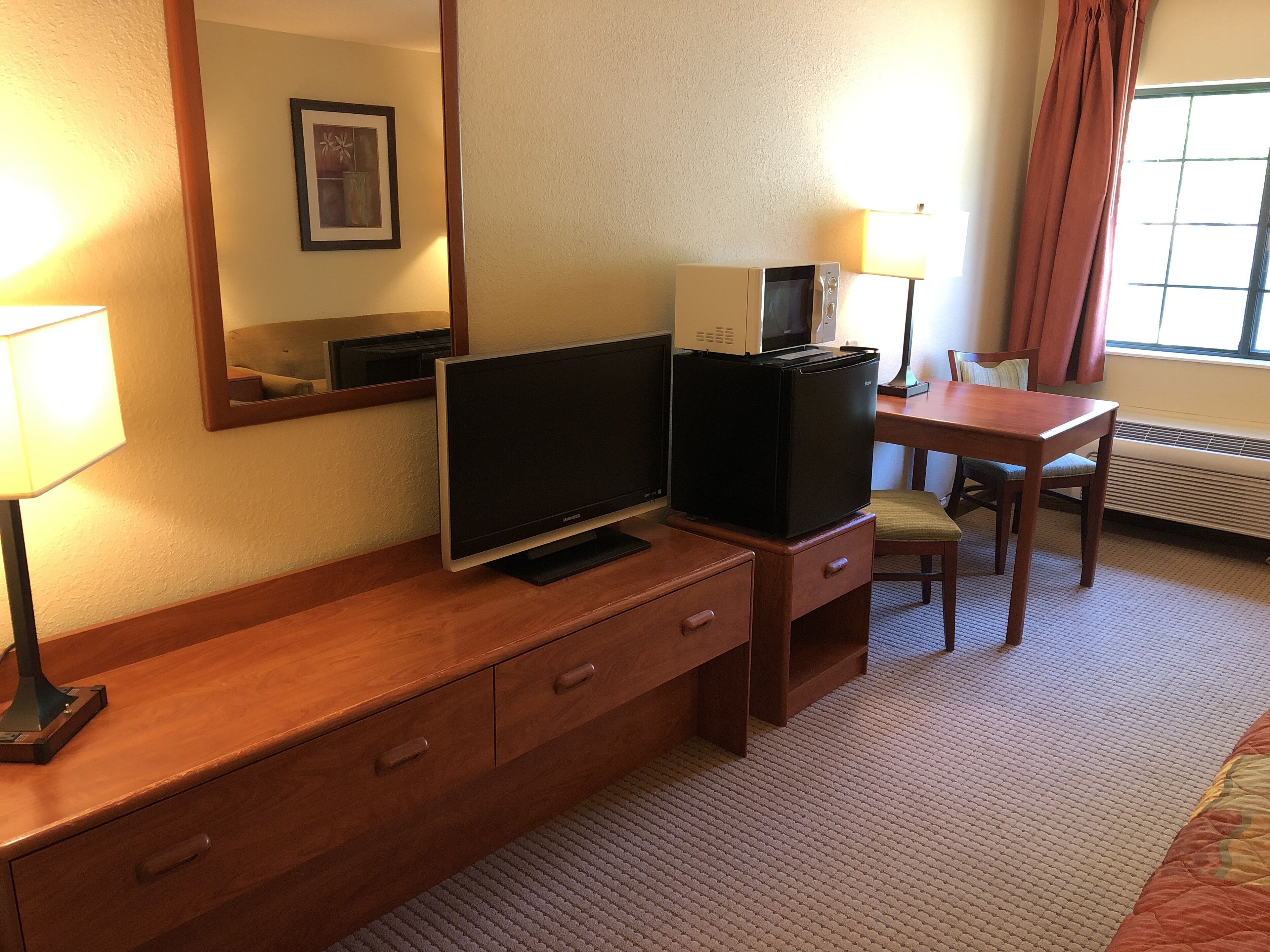 A hotel room with a wooden desk, a television, a small refrigerator, a chair, a lamp, and a large mirror.