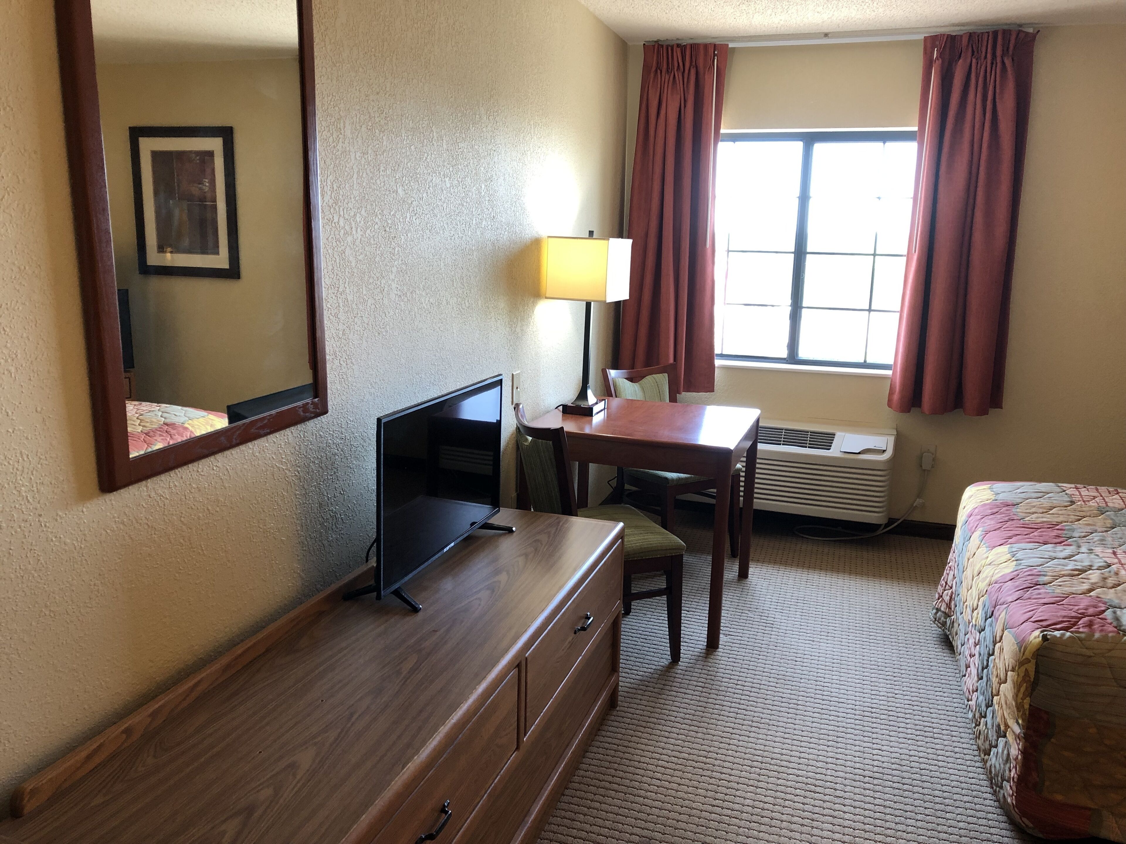 A hotel room with a wooden dresser, a desk with a chair, a bed with a colorful quilt, a window with curtains, and an air conditioning unit.