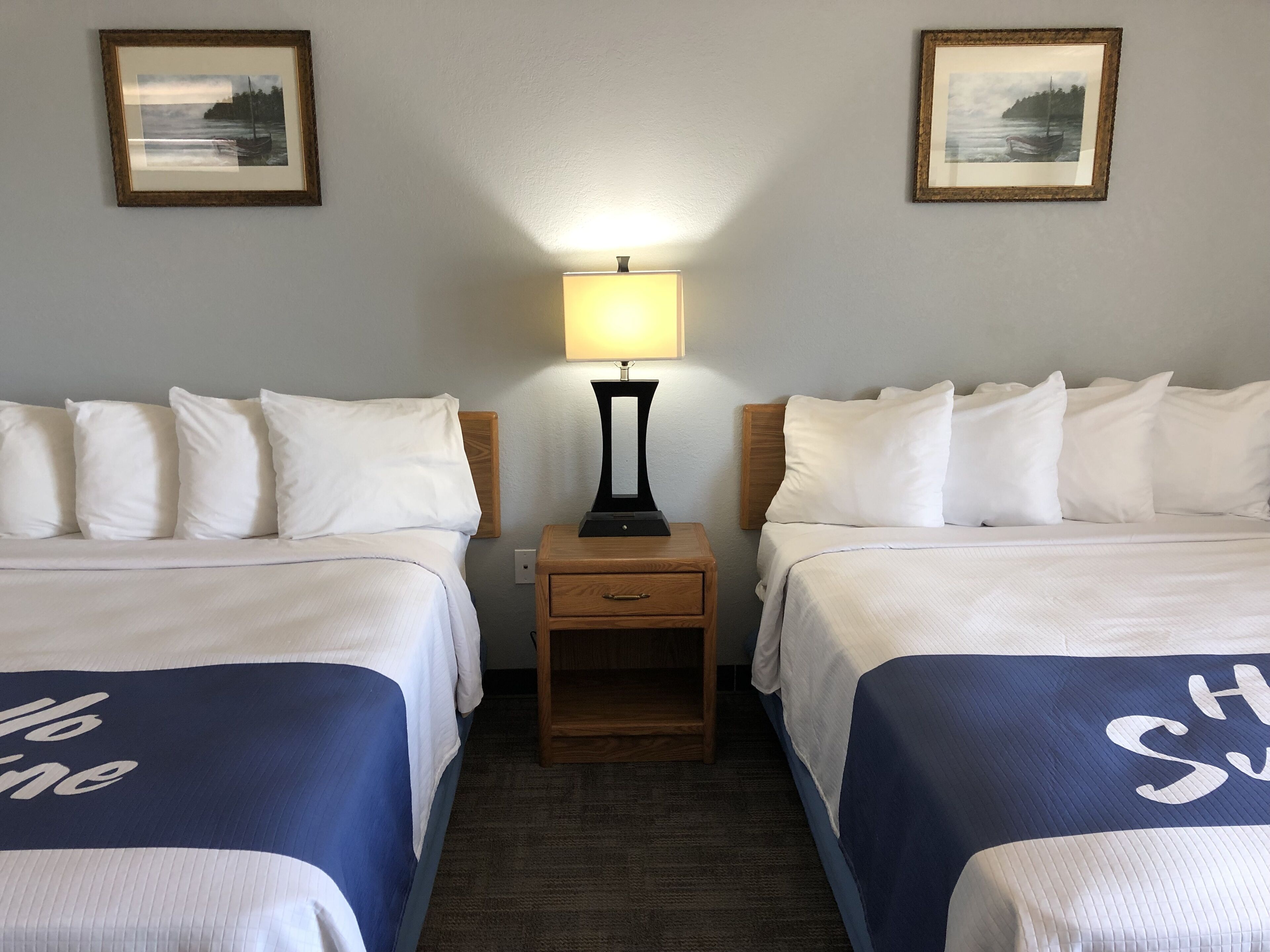 A hotel room with two beds, a nightstand with a lamp, and two framed pictures on the wall.