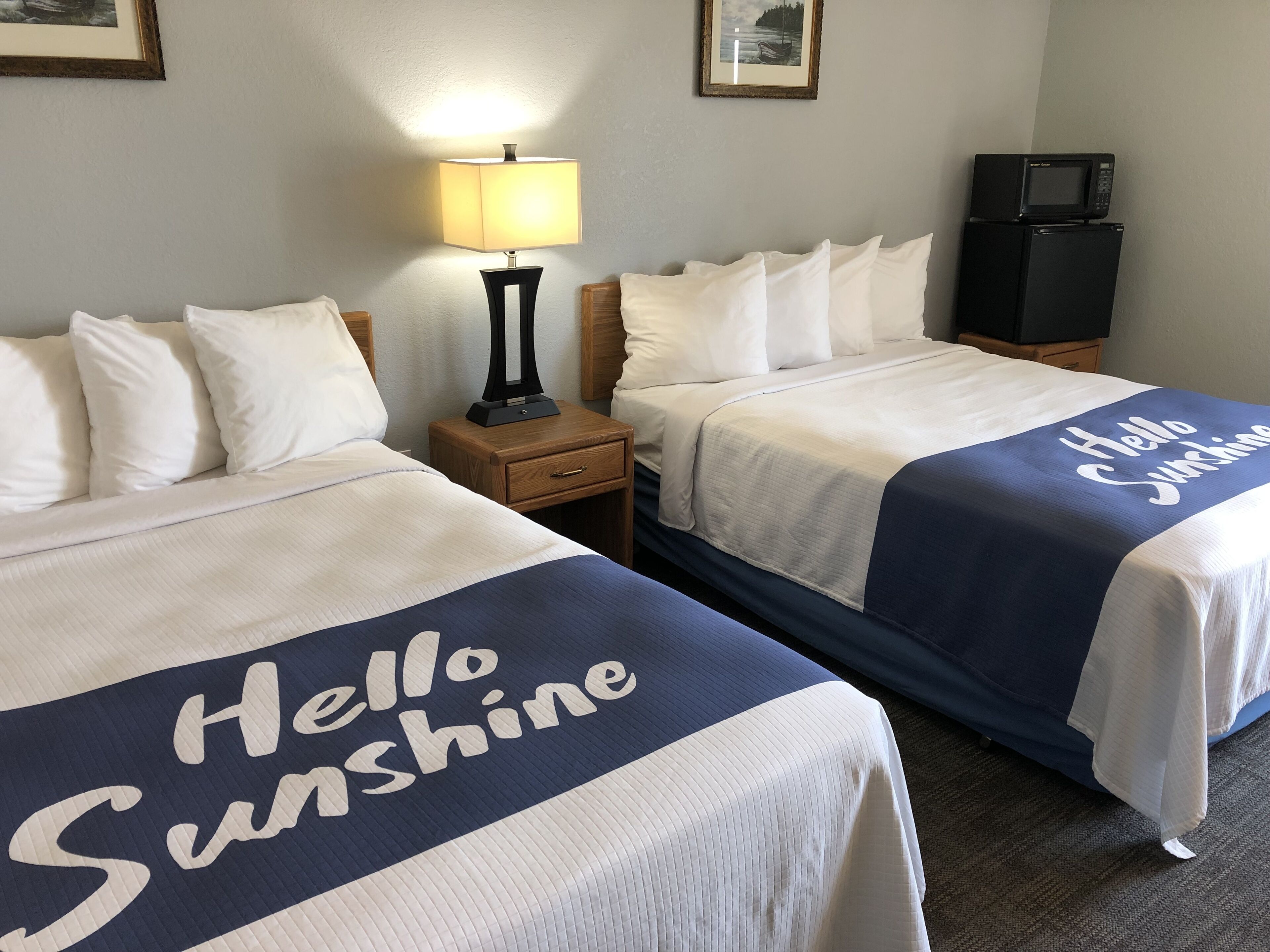 Habitación de hotel con dos camas, cada una con una colcha que dice 'Hello Sunshine'. Hay una mesita de noche con una lámpara entre las camas, y un televisor sobre un soporte a la derecha.