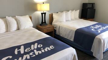 Habitación de hotel con dos camas, cada una con una colcha que dice 'Hello Sunshine'. Hay una mesita de noche con una lámpara entre las camas, y un televisor sobre un soporte a la derecha.