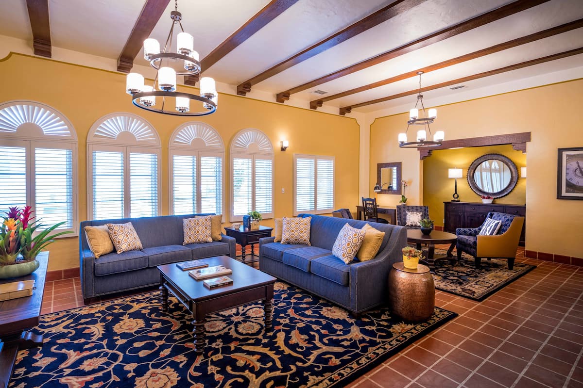 Amplia sala de estar con un conjunto de sofá azul, una mesa de centro y un tapete estampado. Hay varias ventanas con persianas, un candelabro y un comedor con mesa y sillas.