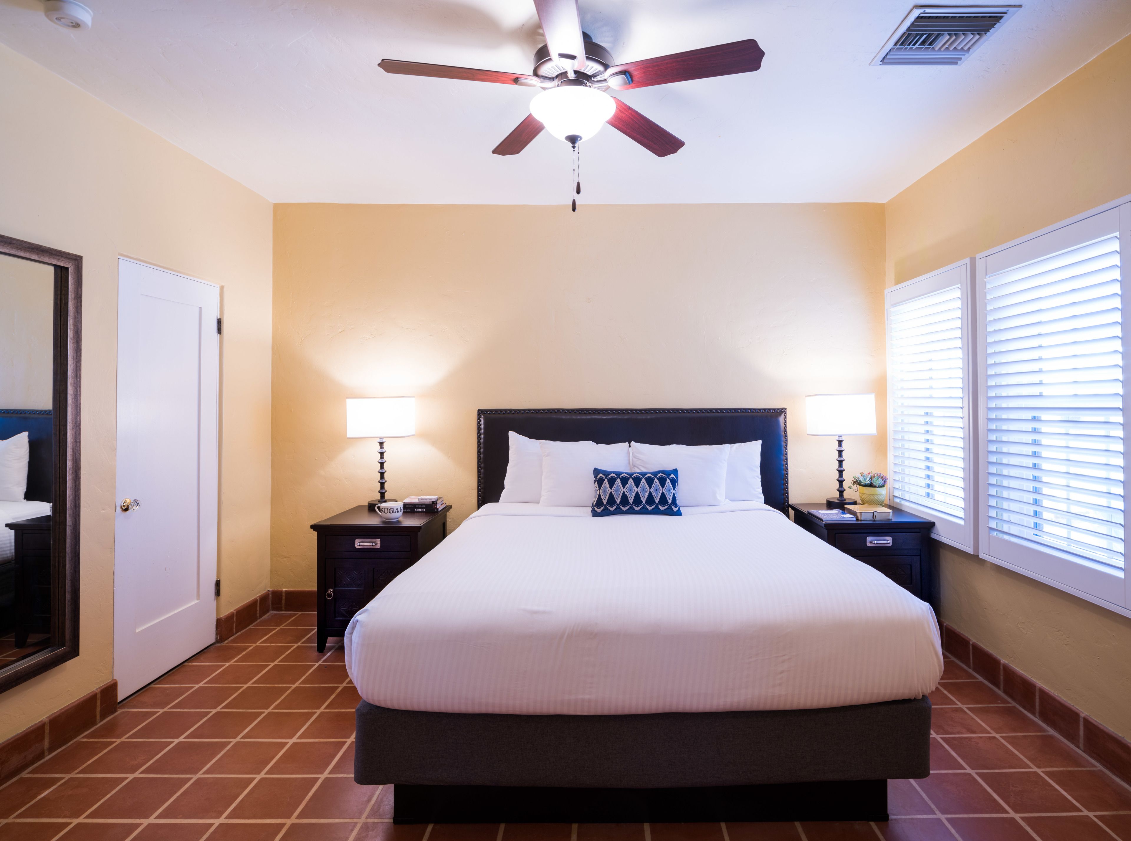 A hotel room with a large bed, two bedside tables with lamps, a ceiling fan, and a window with blinds.