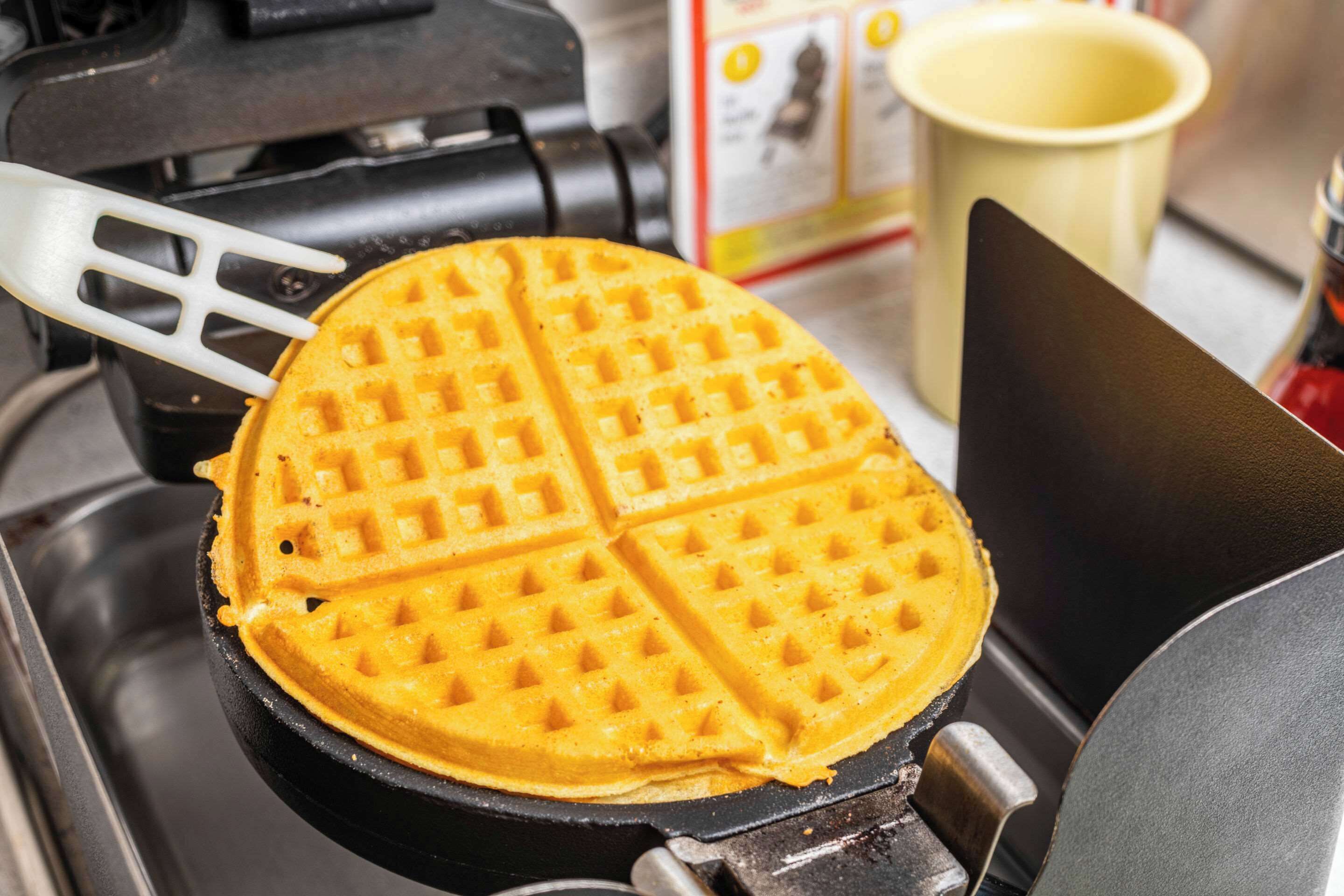 A waffle on a griddle with a spatula and a yellow cup in the background.
