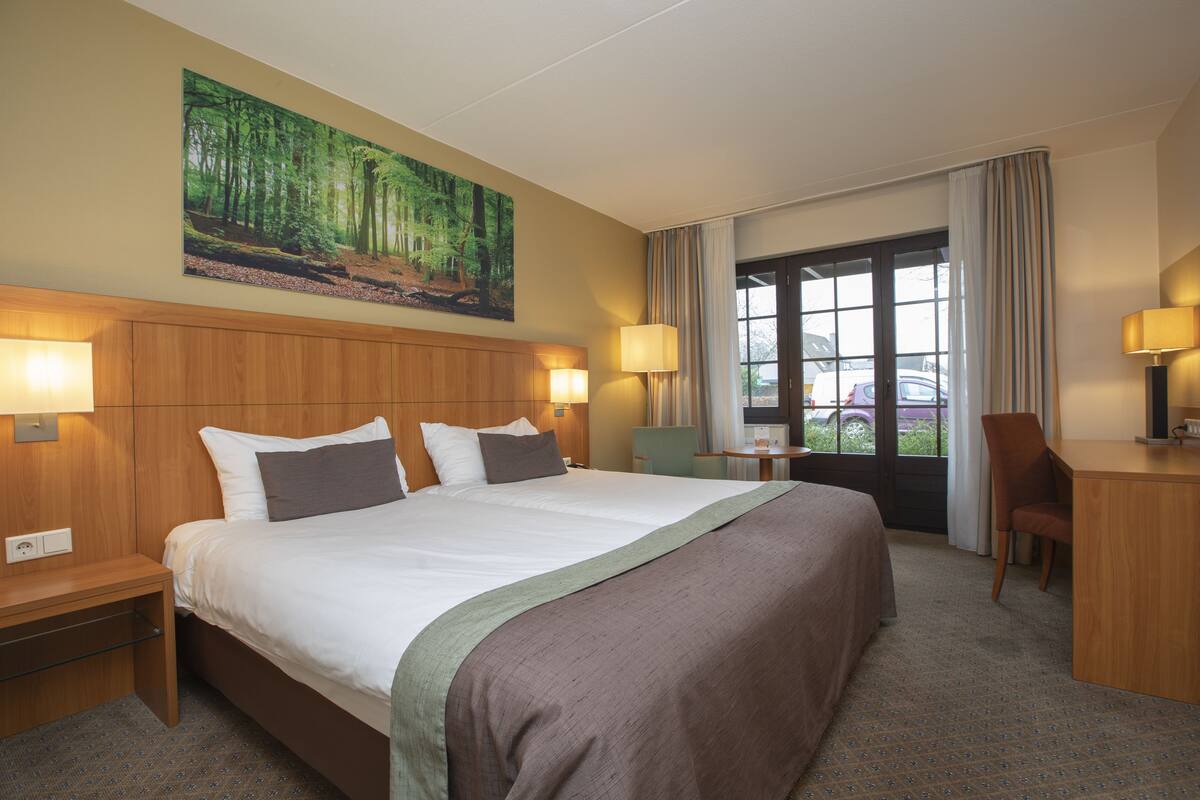 Quarto de hotel com cama grande, duas luminárias de cabeceira, escrivaninha e uma cadeira. Há uma janela com cortinas e um quadro de uma floresta na parede.