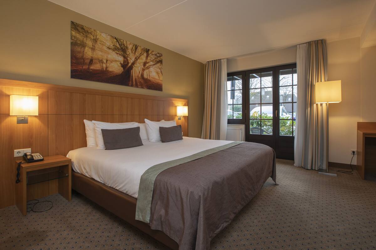 Quarto de hotel com uma cama grande, duas luminárias de cabeceira, mesa de cabeceira com telefone, janela com cortinas e um quadro na parede.