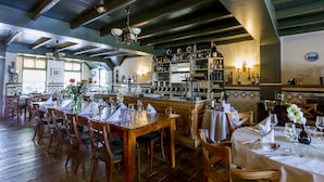 A restaurant with wooden tables and chairs, a bar area with shelves of bottles, and a view of the kitchen through a window. - Fletcher Hotel - Restaurant De Klepperman (Hoevelaken)