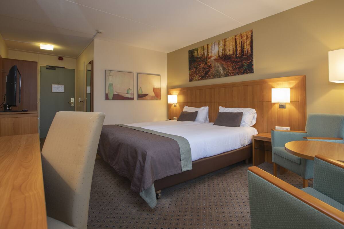Quarto de hotel com cama, duas luminárias de cabeceira, escrivaninha, cadeira e um quadro na parede.