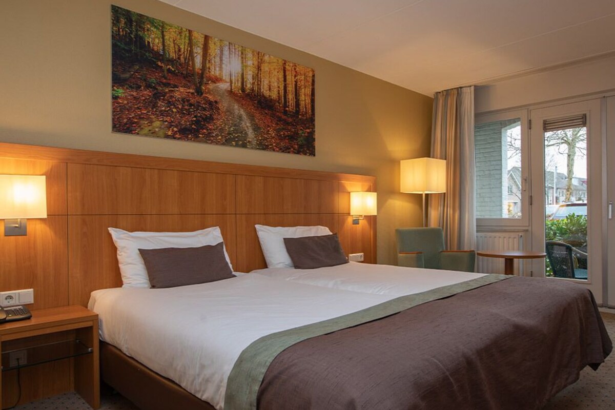 A hotel room with a large bed, two bedside lamps, a painting of a forest path, a window with curtains, and a small table with a chair.