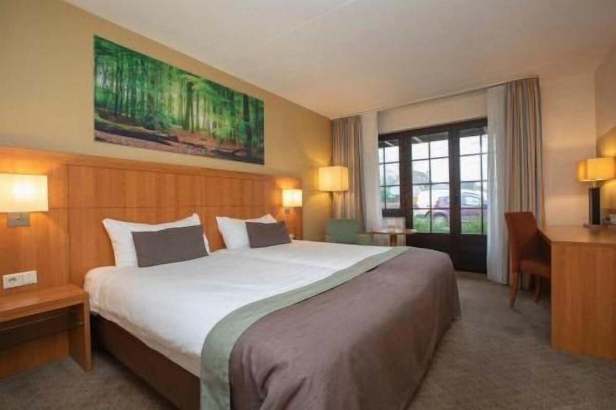 A hotel room with a large bed, two bedside lamps, a desk with a chair, a painting on the wall, and a window with curtains.