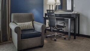 A hotel room with a grey armchair, a desk with a lamp, and a large mirror.
