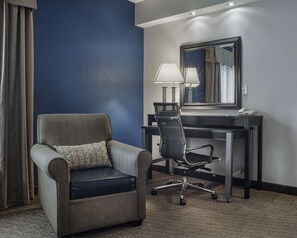 Una habitación de hotel con un sillón gris, un escritorio con lámpara y un espejo grande.