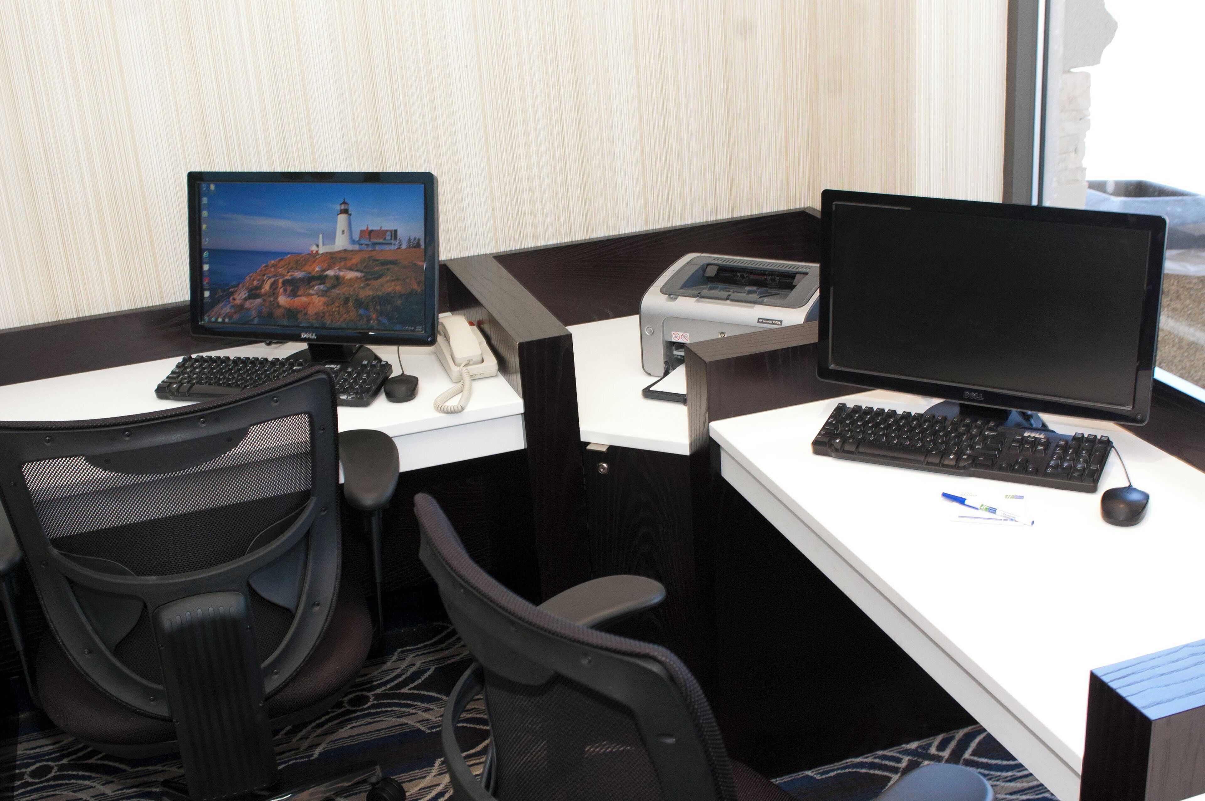 A hotel room with two desks, each with a computer monitor, keyboard, and mouse. There is a telephone on one desk and a printer on the other. The room has a carpeted floor and a window with blinds.
