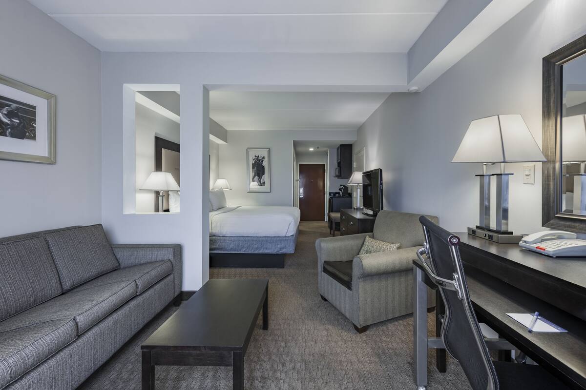 A hotel room with a sofa, a bed, a desk with a chair, and a desk lamp.