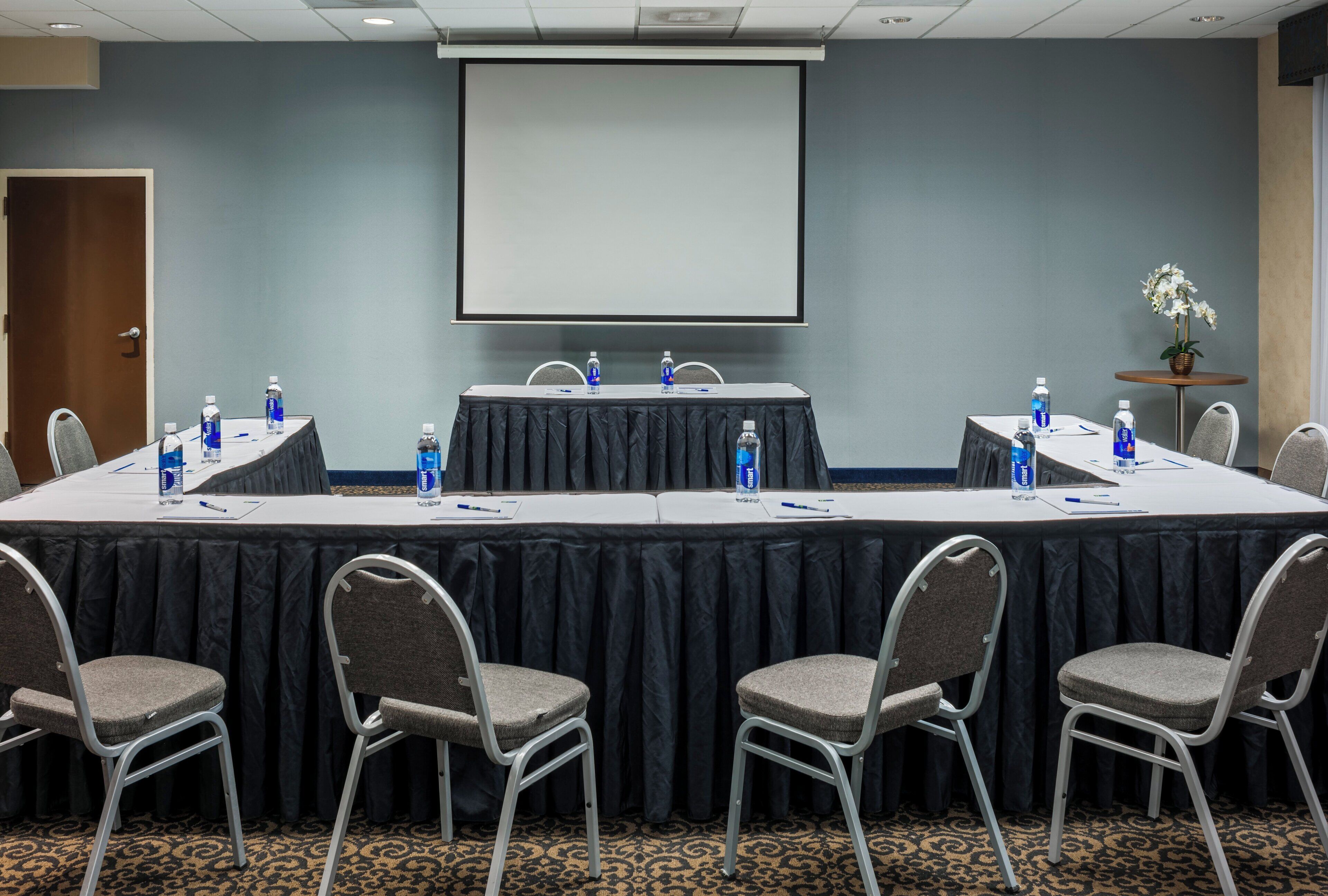 Un salón de conferencias con una larga mesa, sillas, una pantalla de proyector y botellas de agua.