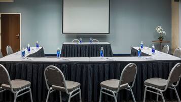 Un salón de conferencias con una larga mesa, sillas, una pantalla de proyector y botellas de agua.