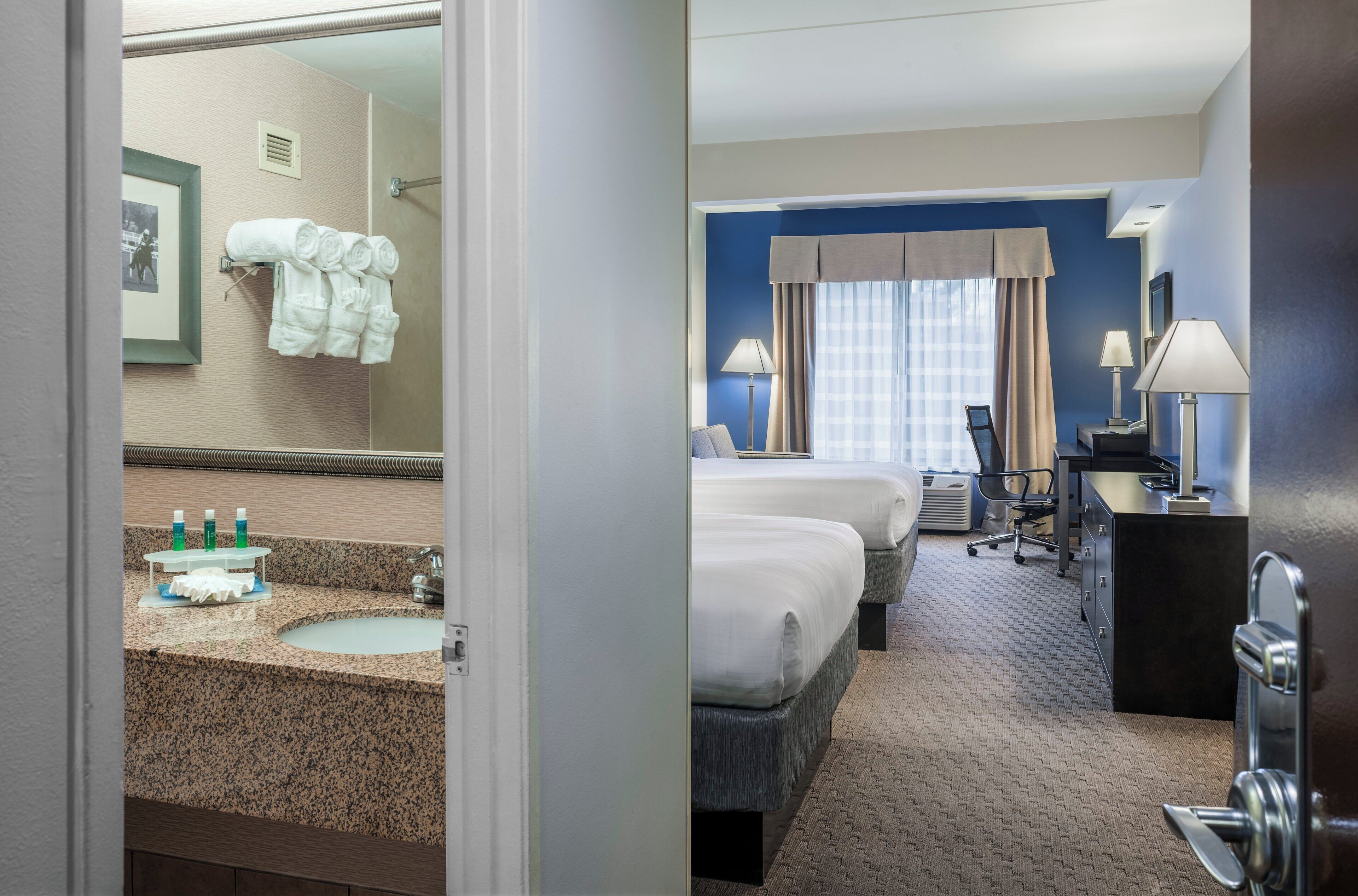 A hotel room with a bathroom, two beds, a desk, and a chair.