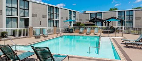 Una piscina rectangular con sillones de descanso y sombrillas, rodeada por una cerca blanca y un edificio con amplias ventanas.