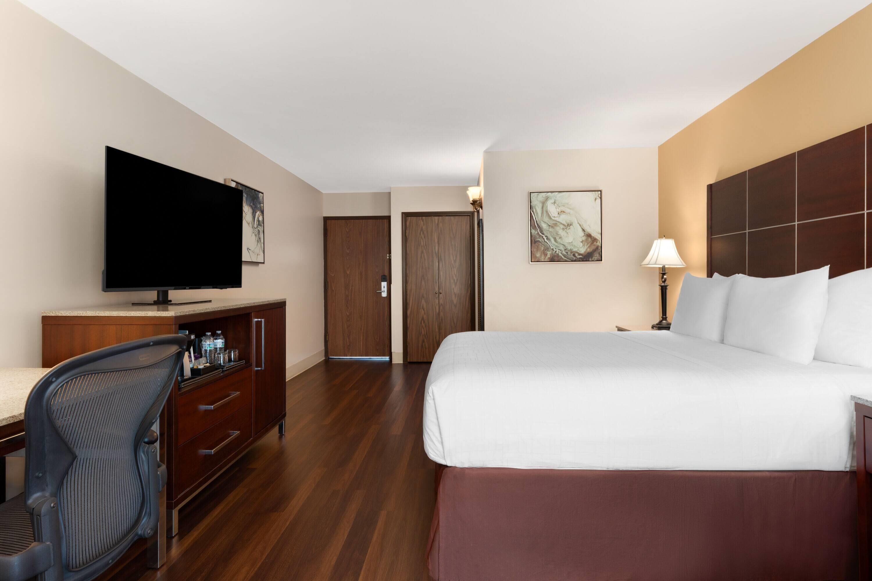 A hotel room with a large bed, a desk with a chair, a television, and a door leading to another room.