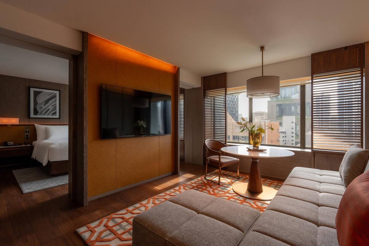 Une chambre d’hôtel moderne équipée d’un canapé, d’une table à manger avec des chaises, d’un téléviseur fixé au mur et de grandes fenêtres avec des stores.