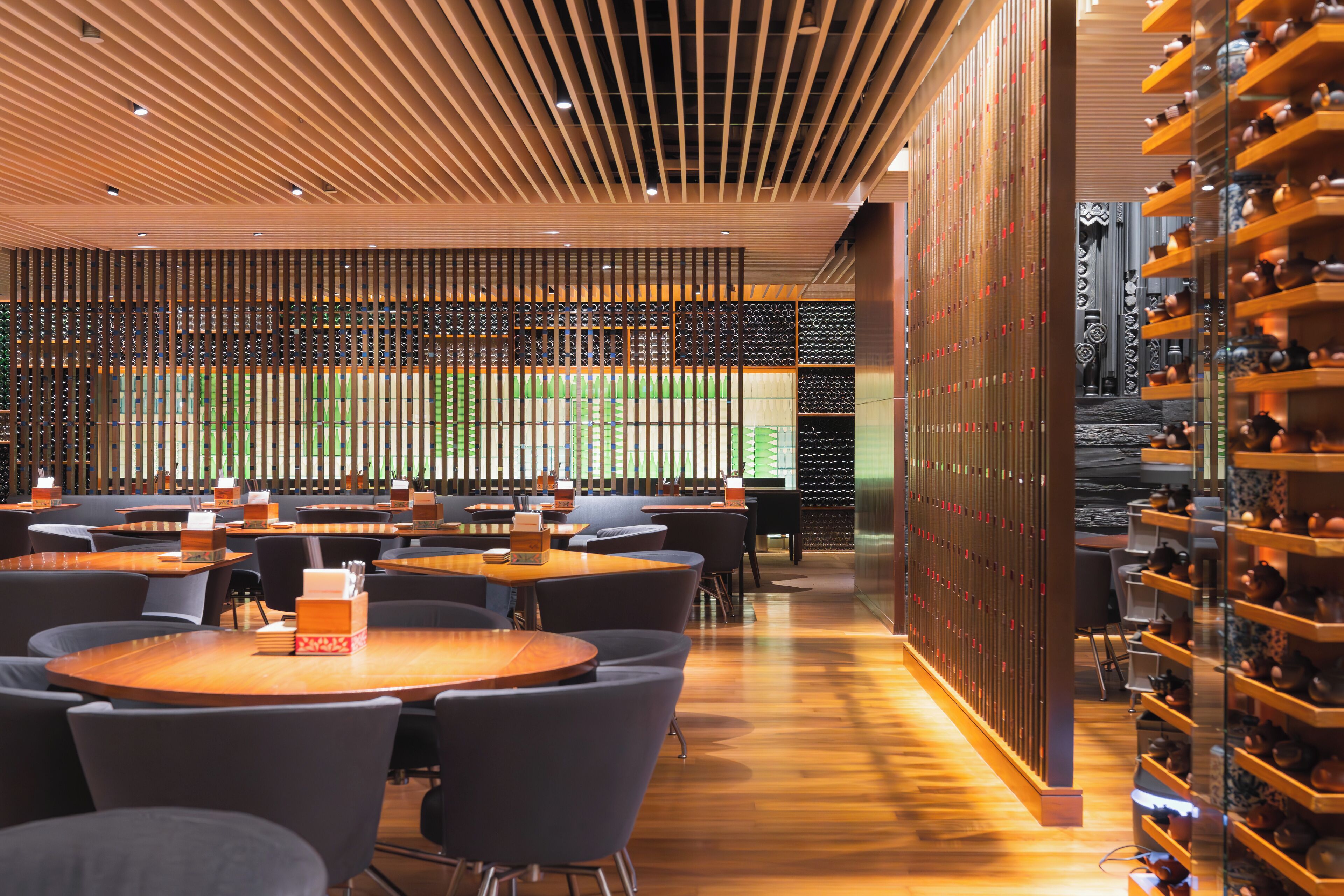 A modern restaurant interior with wooden tables and chairs, a decorative wall with vertical slats, and a wine rack on the right.