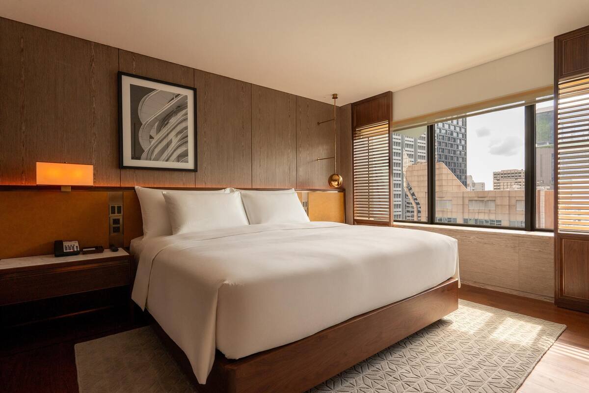 Une chambre d’hôtel moderne avec un grand lit, une tête de lit en bois, un tableau encadré, une lampe de chevet et une vue sur la ville grâce à de grandes fenêtres.