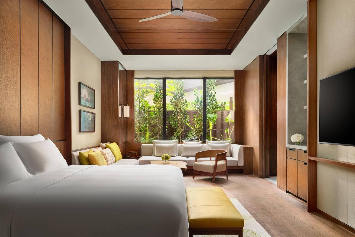 Une chambre moderne avec un grand lit, un coin salon avec des fauteuils, une télévision et une vue sur la verdure environnante.
