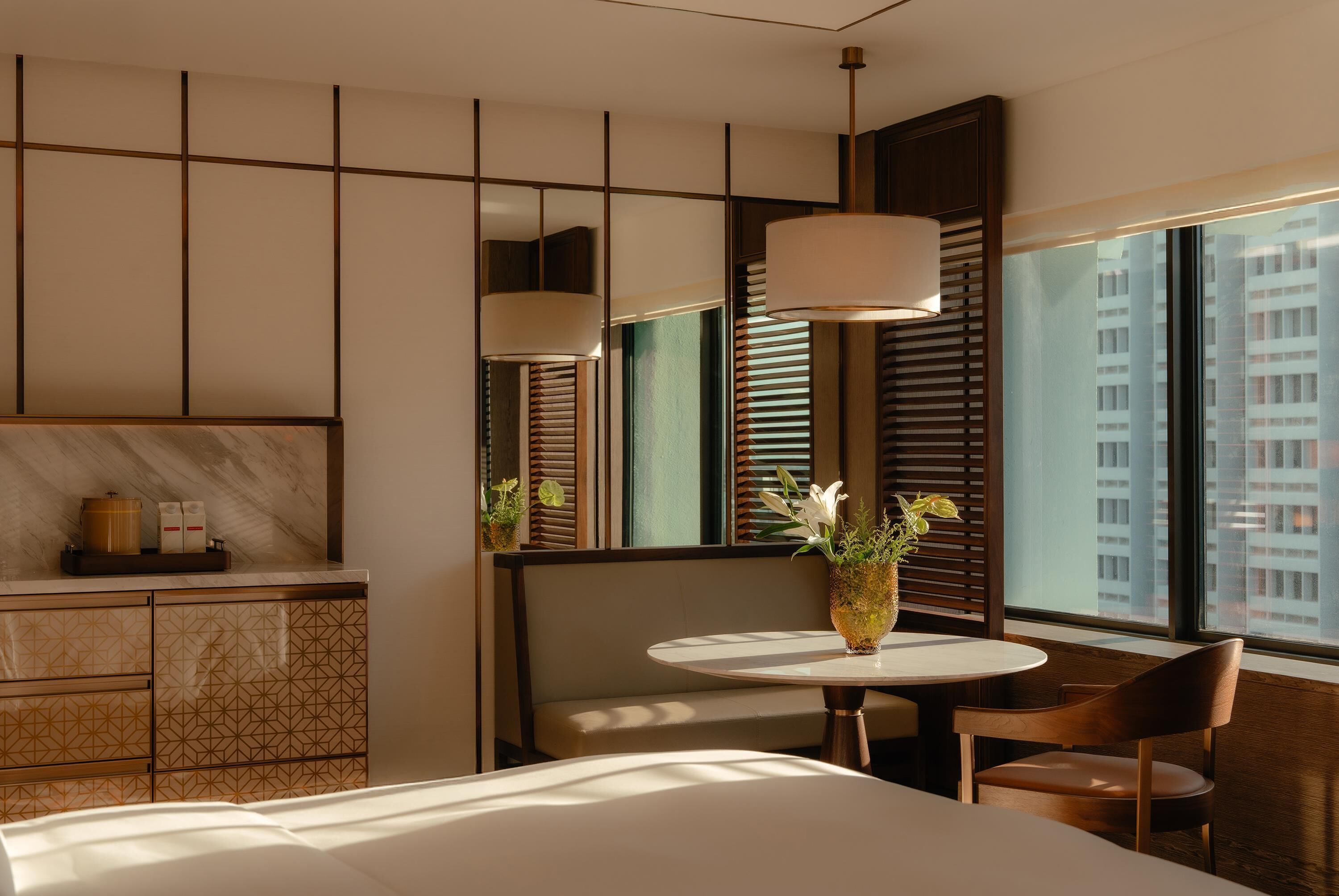 Une chambre d’hôtel avec un lit, une table ronde ornée d’un vase de fleurs, un banc et une vue sur de hauts immeubles, visible à travers de grandes fenêtres.