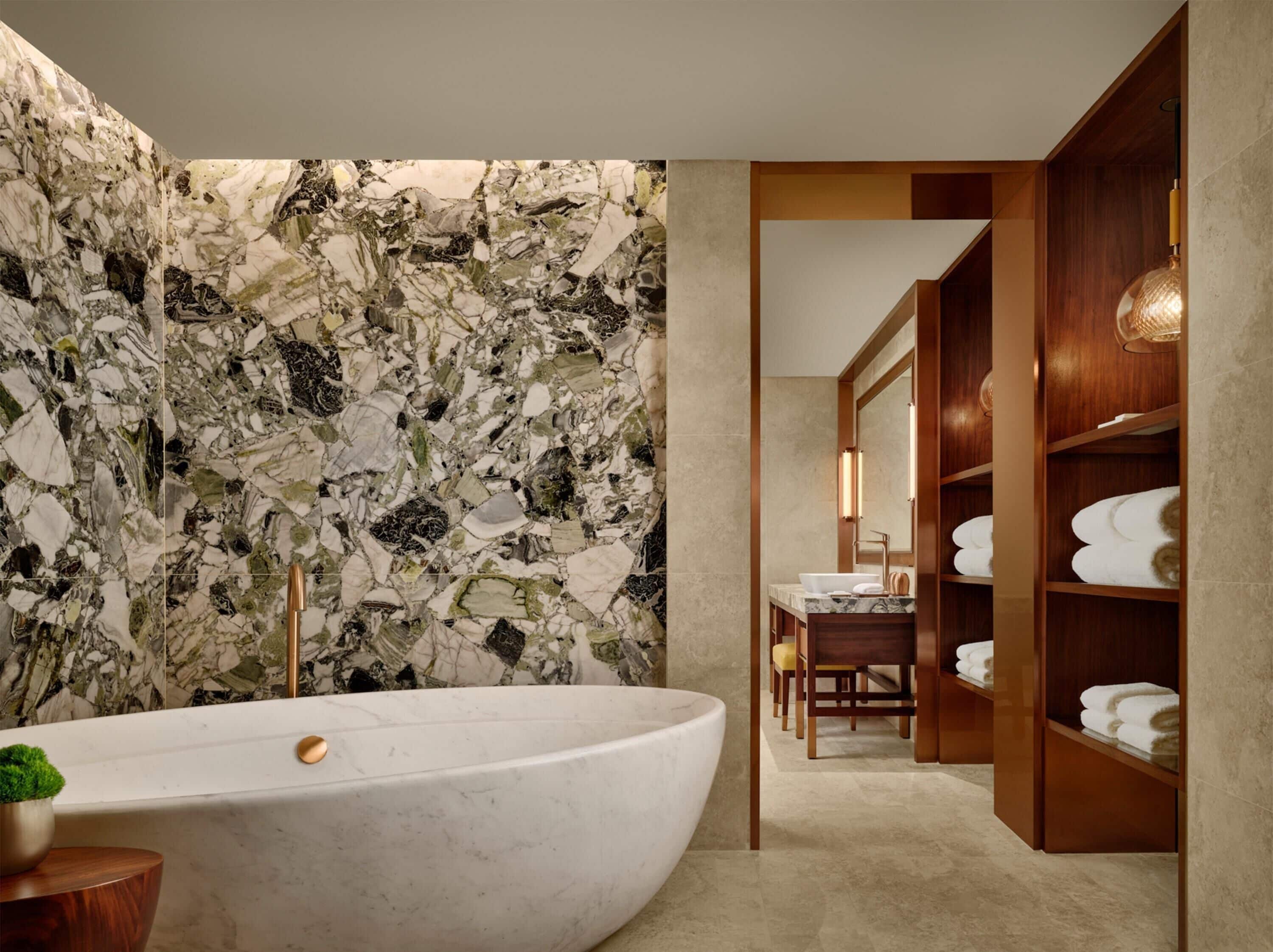 A modern bathroom with a large freestanding bathtub, a marble wall, and a wooden vanity with a mirror and shelves for towels.