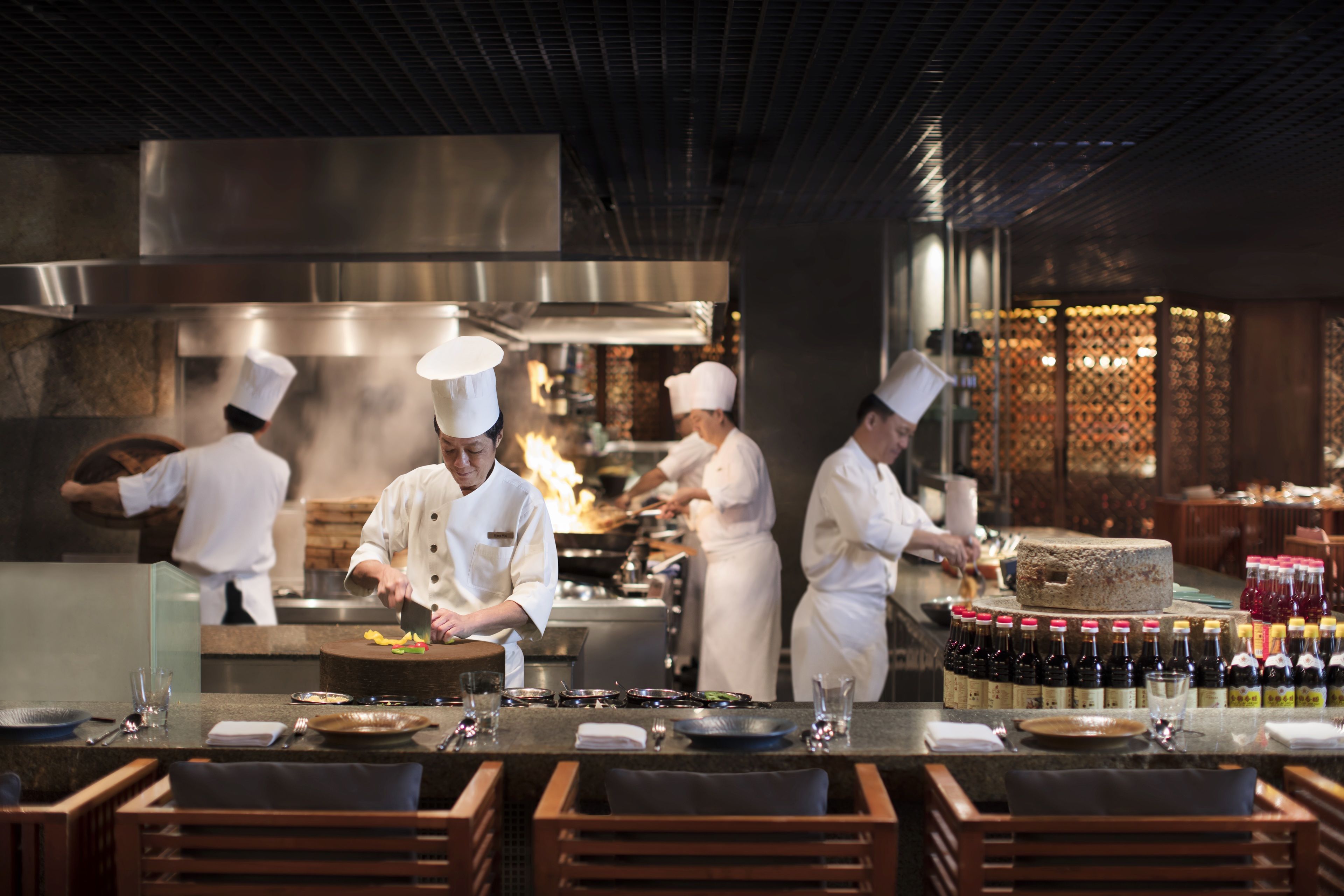 A professional kitchen with chefs preparing food, a variety of bottles on the counter, and wooden chairs in the foreground.