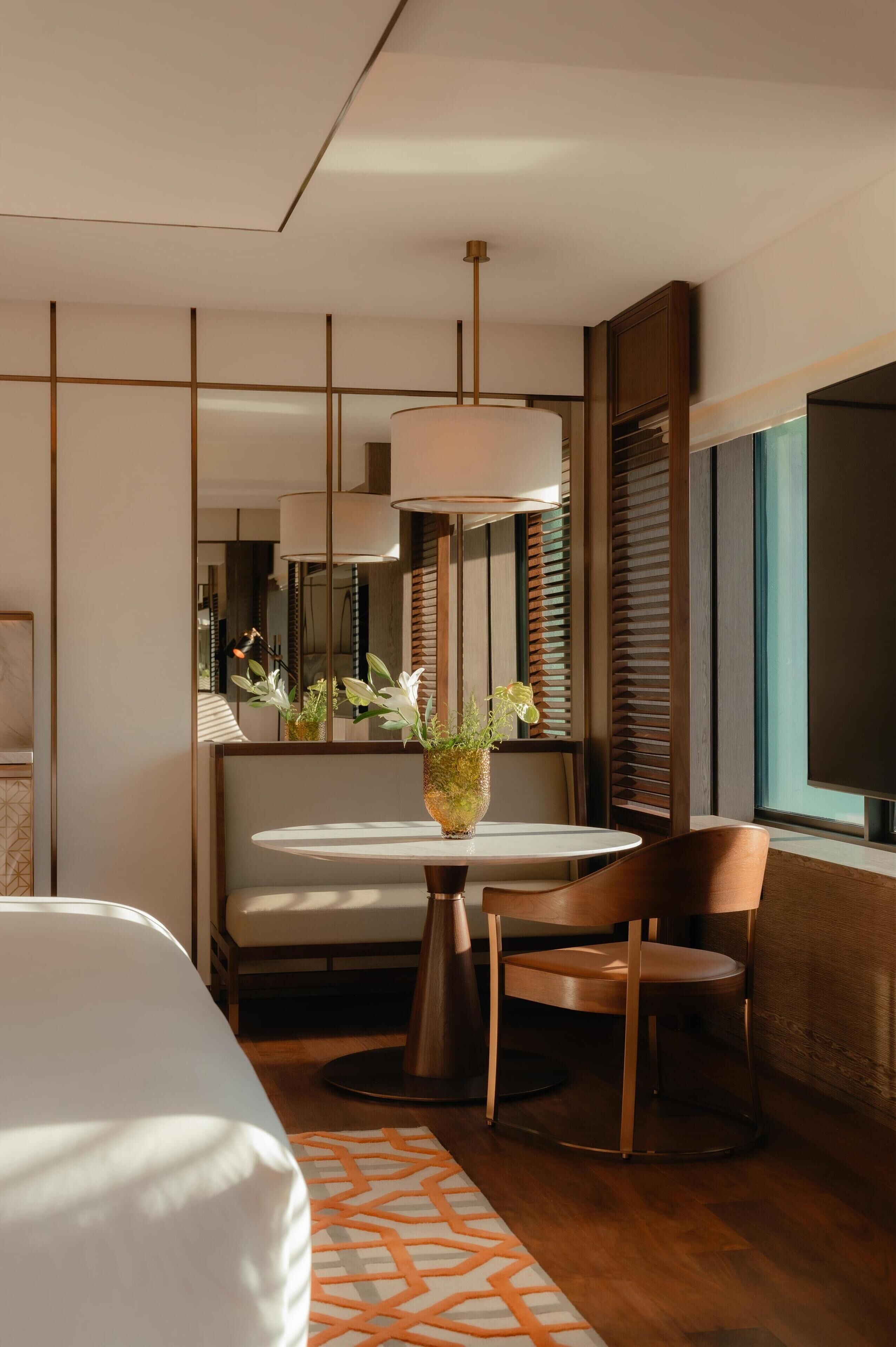 A modern hotel room with a bed, a round table with chairs, a vase with flowers, and a large window.
