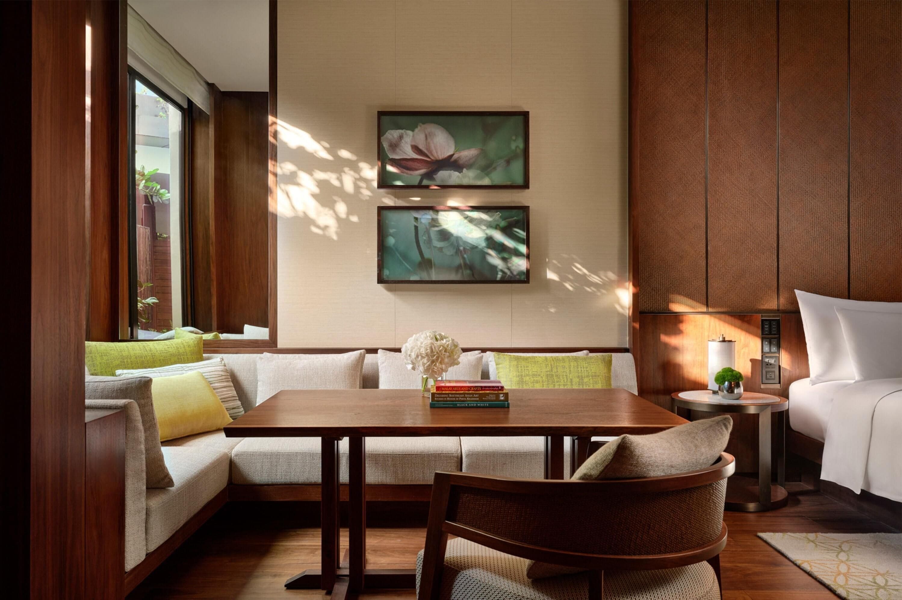 Een moderne woonkamer met een houten tafel, bank en fauteuil. Twee ingelijste foto’s hangen aan de muur, een klein bijzettafeltje met een lamp en een raam met zonwering.