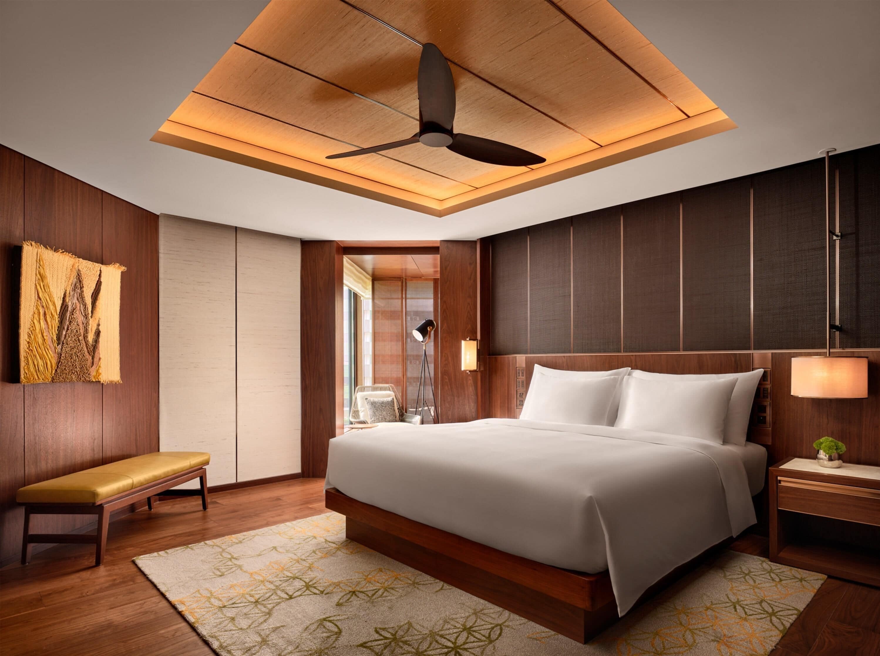 A modern hotel room with a large bed, wooden paneling, a ceiling fan, a bench, and a bedside table with a lamp.
