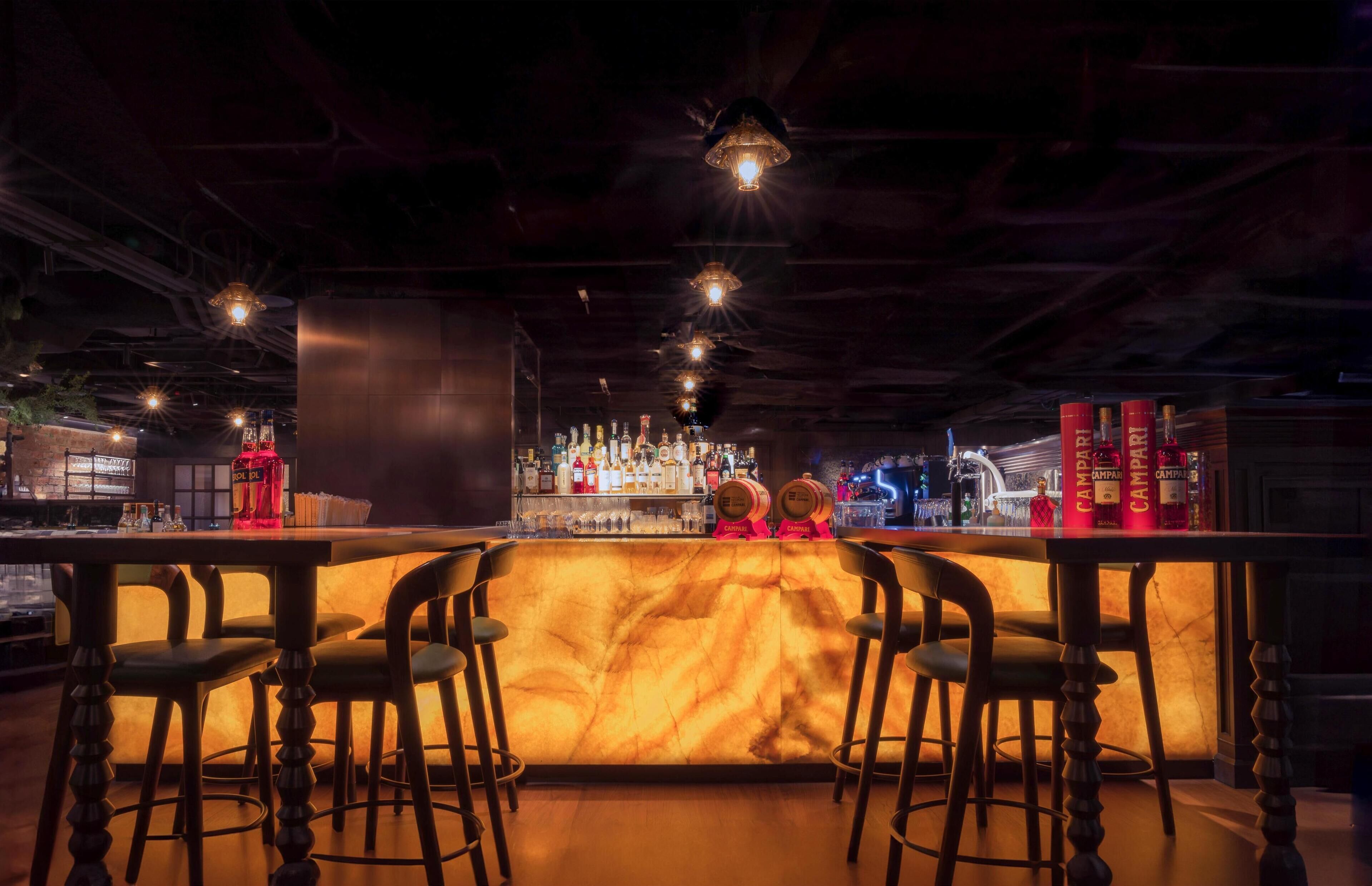 A bar area with a marble countertop, multiple stools, and a well-stocked bar with various bottles and a red bottle prominently displayed.