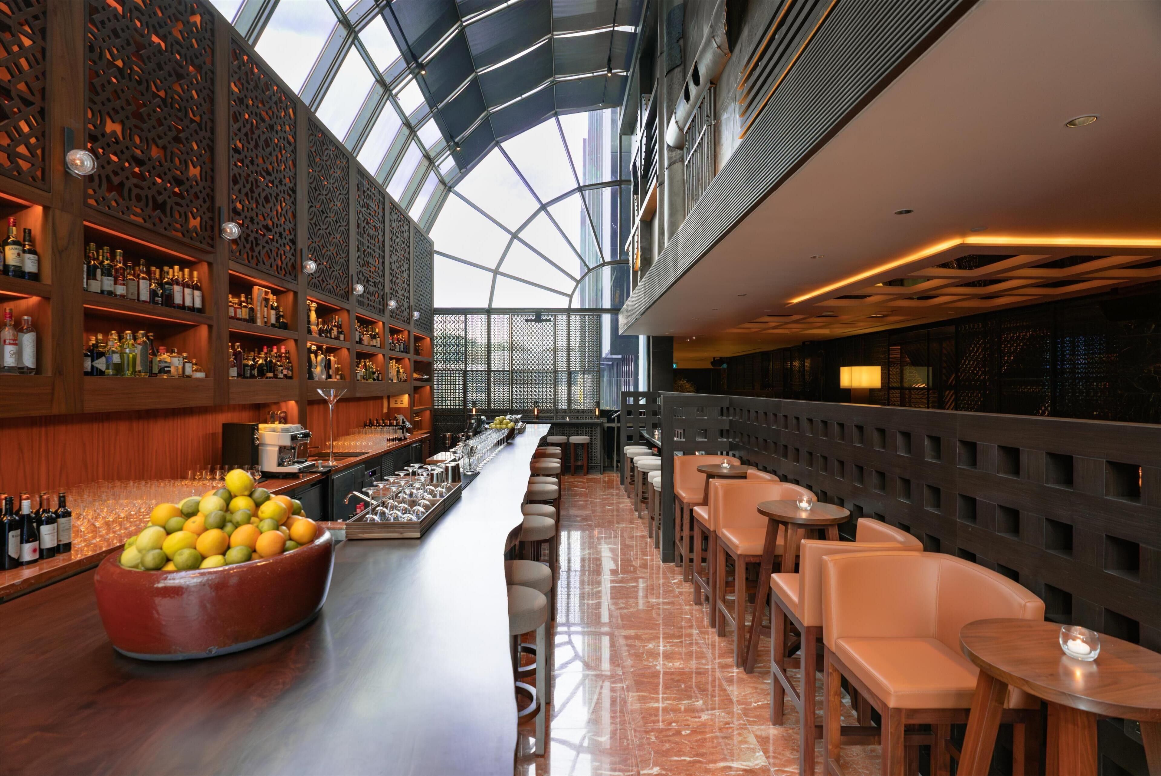 A modern bar with a high ceiling, wooden shelves with bottles, a large fruit bowl, and seating areas.