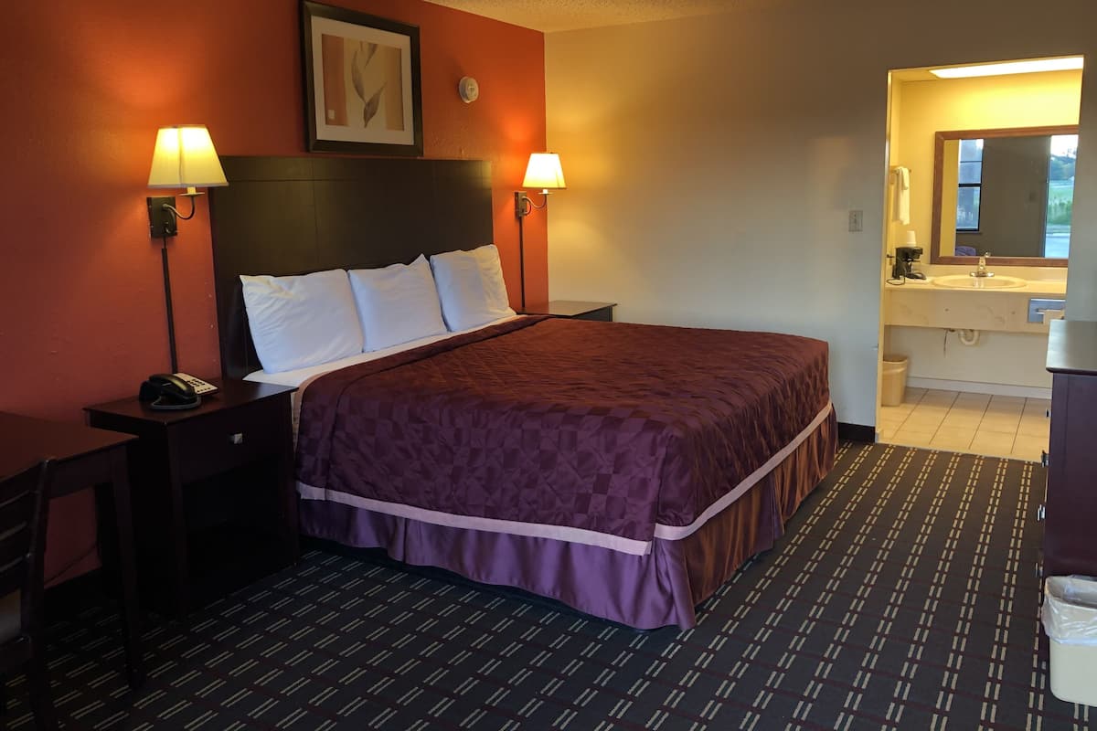 Habitación de hotel con una cama grande, dos lámparas de noche, una mesita de noche con un teléfono, un cuadro en la pared y un baño visible a través de una puerta abierta.
