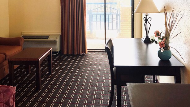 A hotel room with a sofa, a small table, a dining table with a lamp and flowers, a large window with curtains, and a radiator. - Executive Inn & Suites (Prescott)
