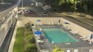 Une piscine entourée d’une clôture, avec des chaises longues et des parasols, un parking et un bâtiment en arrière-plan.