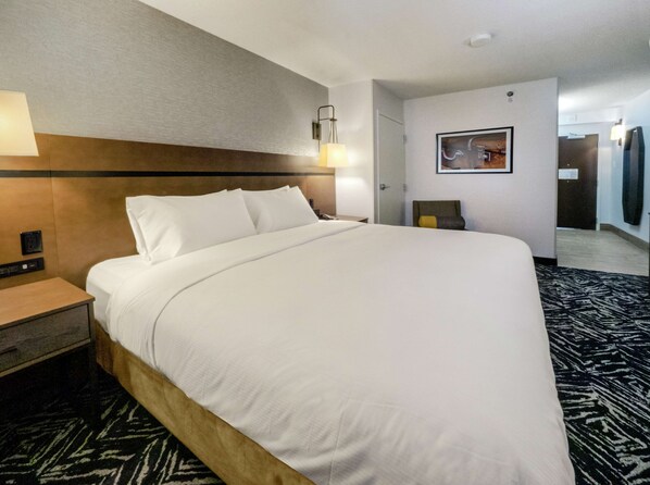 Habitación de hotel con una cama grande, una mesita de noche, una lámpara de pared, un cuadro y una puerta que da a otra habitación.
