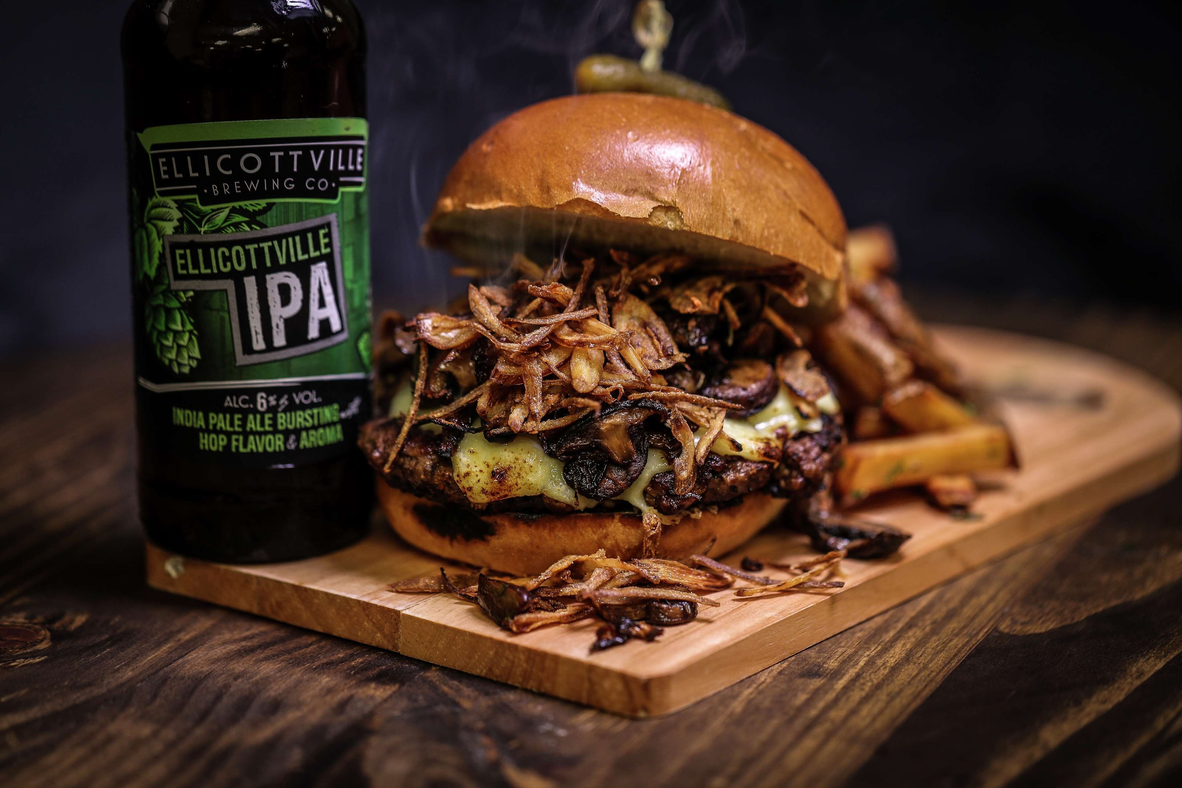 A burger with onions and a bottle of beer on a wooden board.