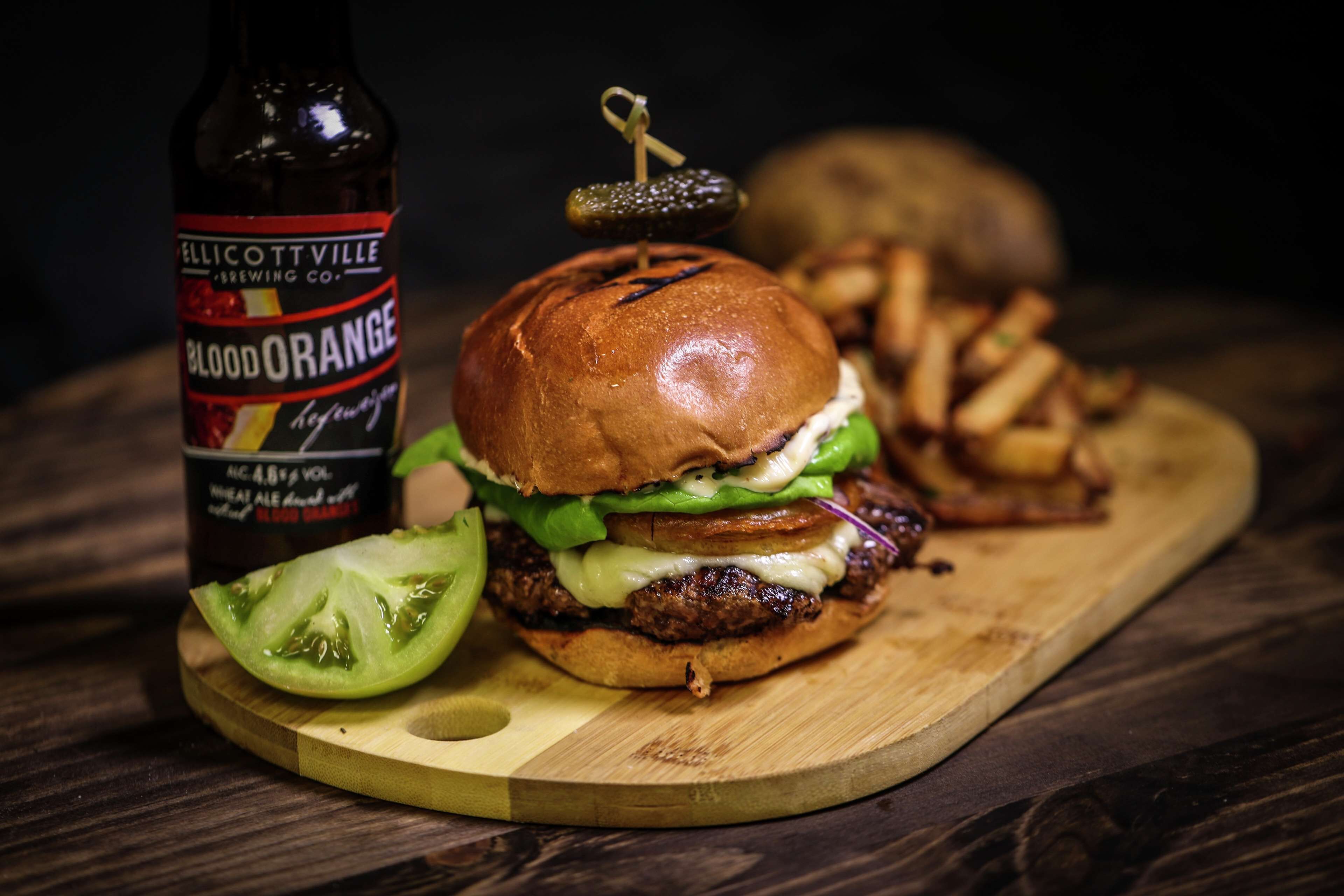 Ein Burger mit einer Gewürzgurke, einer Tomatenscheibe und eine Flasche Bier auf einer Holzbrett.