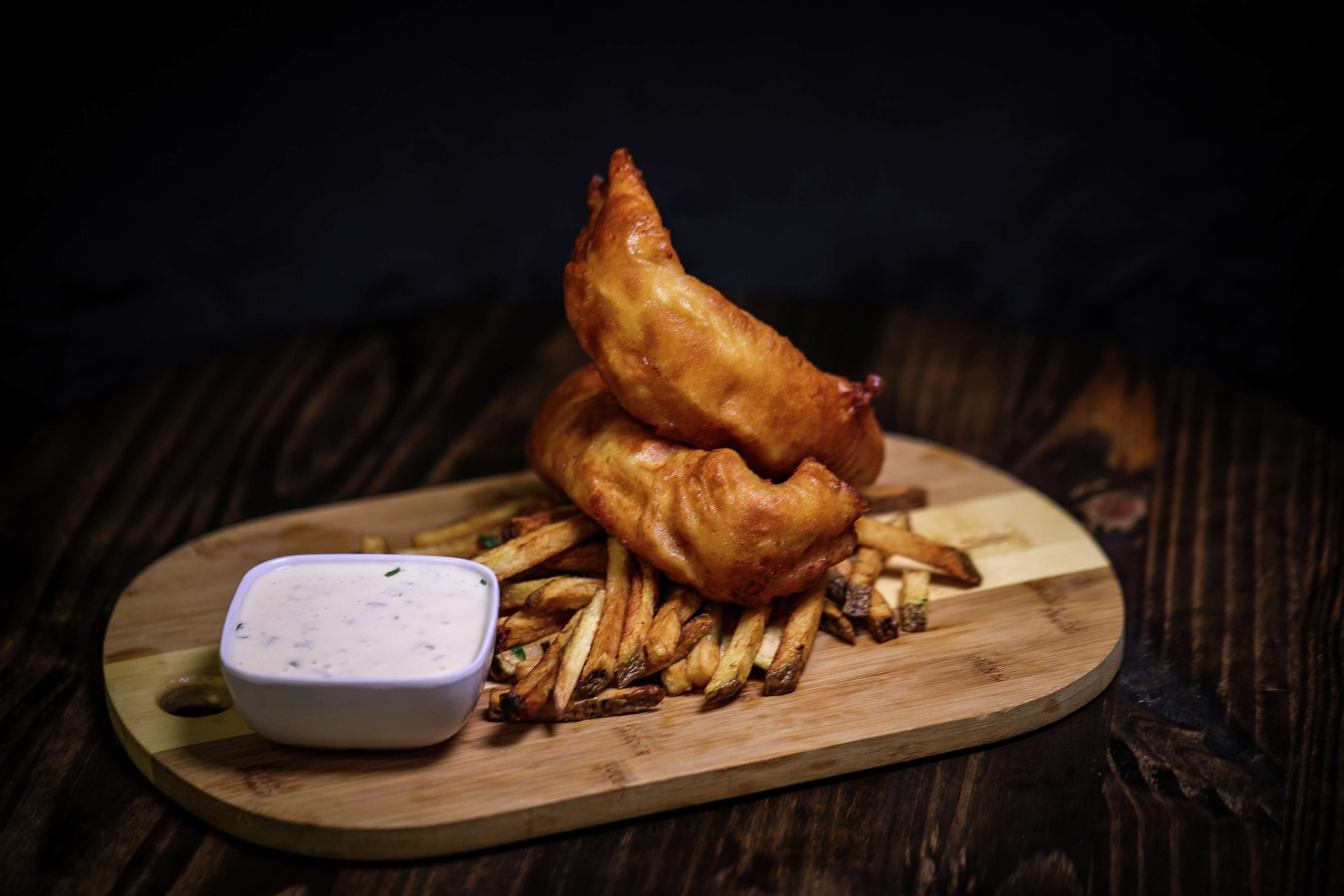 A wooden serving board with a stack of golden-brown fish and a small bowl of white dipping sauce.